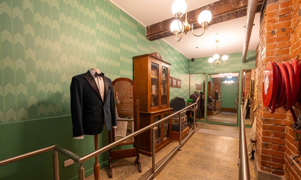 A corridor with an old bookshelf, colourful wall paper and a mannequin wearing a tuxedo.