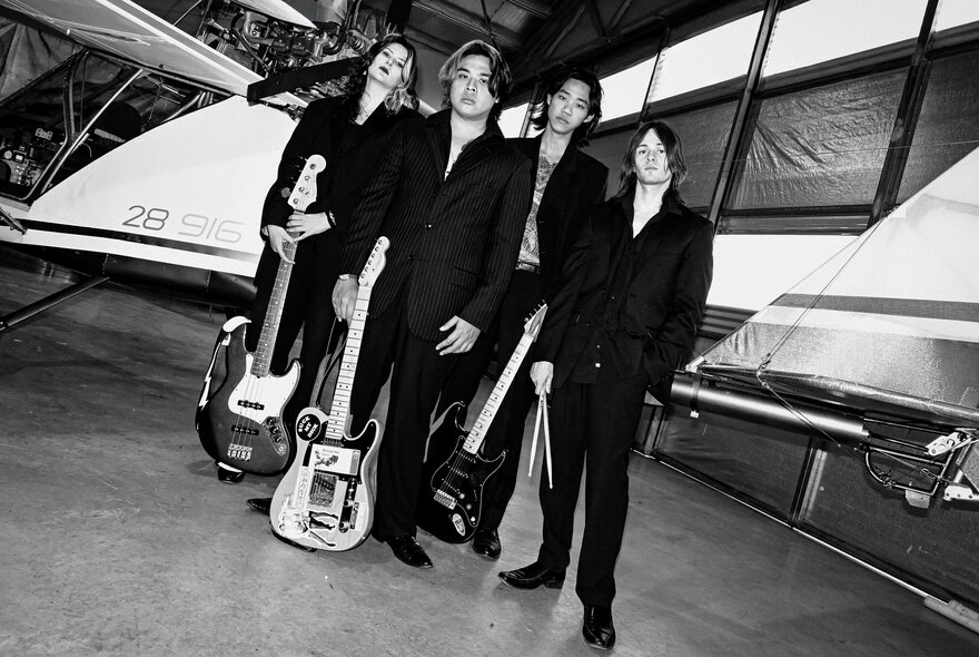Four band members posing, three of them holding their guitars and the fourth holding drumsticks; black and white image.