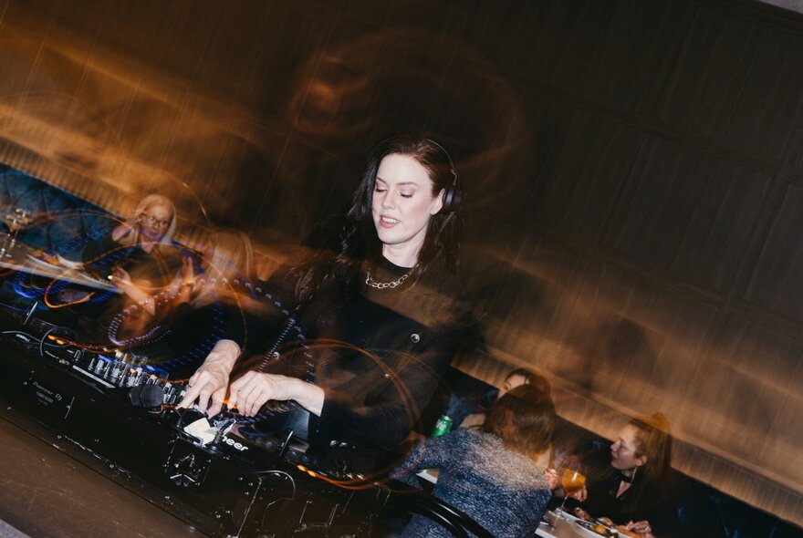 A woman with dark hair stands behind a DJ controller in a dimly lit club or restaurant setting.