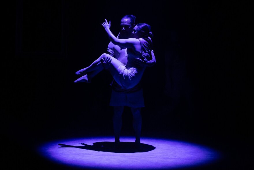 A man standing in the centre of a darkened stage, carrying a person in his arms, both of them under a cool blue spotlight.