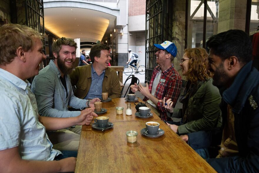 Six people sitting at a table in a cafe with coffees, joking and interacting. 