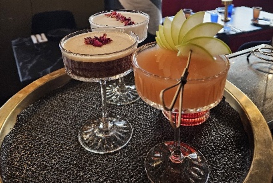 Three cocktails in intricate glassware on a black-lined metal tray.