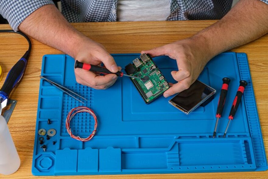 A pair of hands using a small screwdriver to work on an electronic circuit board, with other tools and wire nearby, all resting on a blue worksurface mat.