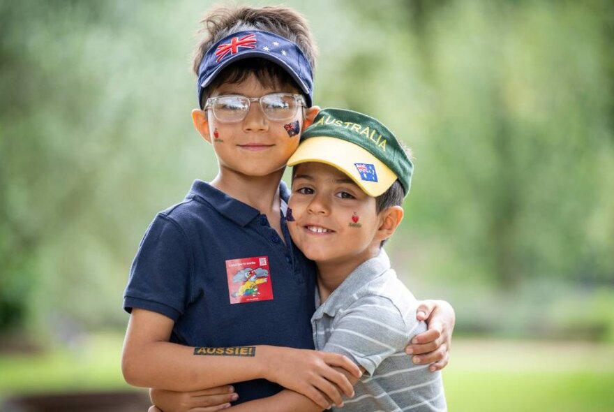 Two children wearing Australia Day hats and face stamps, hugging against greenery.