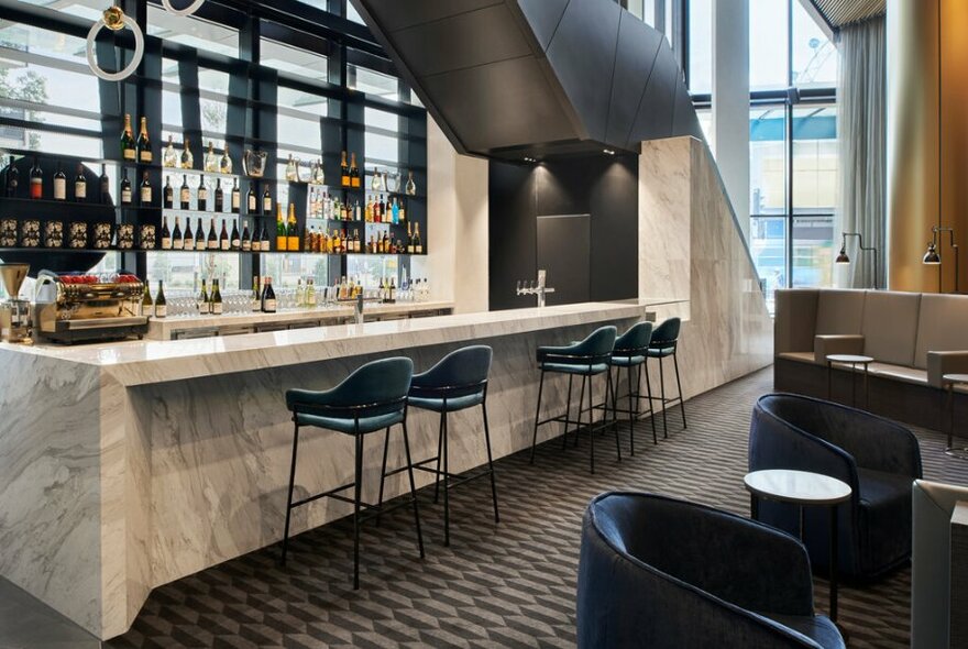 The marble bar at Storehouse restaurant, with bar stools facing into the bar, rows of bottles displayed against a window, and to the right club armchairs and small tables.