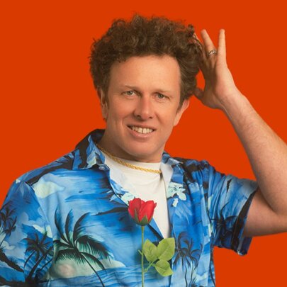 Comedian Nick Capper, a middle-aged man with curly brown hair, wearing a Hawaiian shirt and raising his hand to his head.
