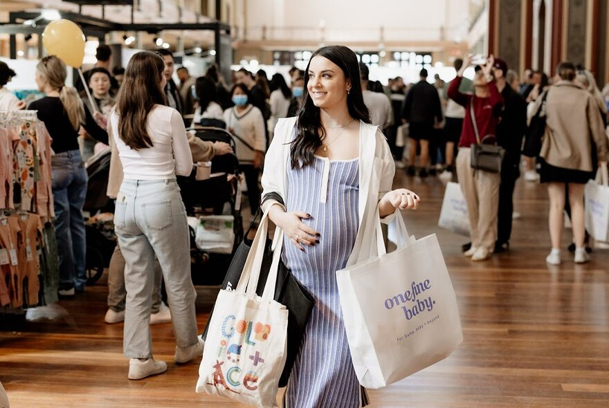 A pregnant woman carrying tote bags over her arms, with her hand resting on her belly, walking through a baby expo in a large hall, with people in the background.