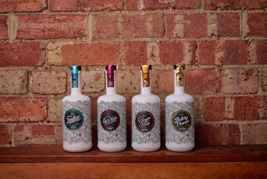 A line up of white bottles of gin, each with a different coloured cap, against a brick wall. 