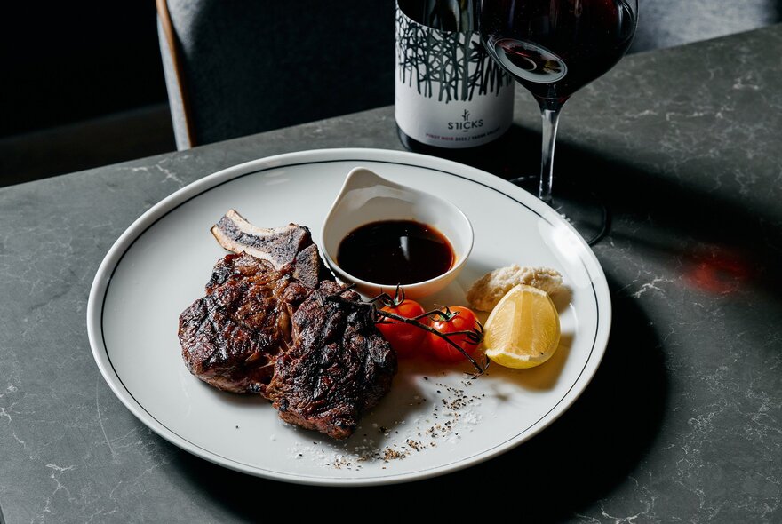 A bone-in steak on a white plate with sauce, cherry tomatoes, and a lemon wedge, next to a glass of red wine.