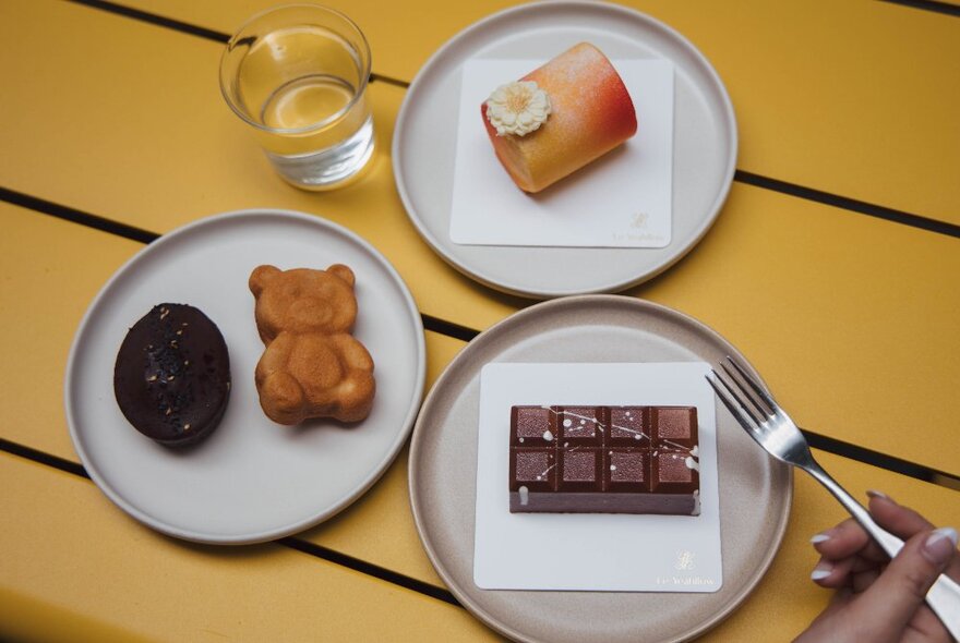 Three plates with sweet treats on a yellow table, including a chocolate cake that has a hand holding a fork hovering over it.