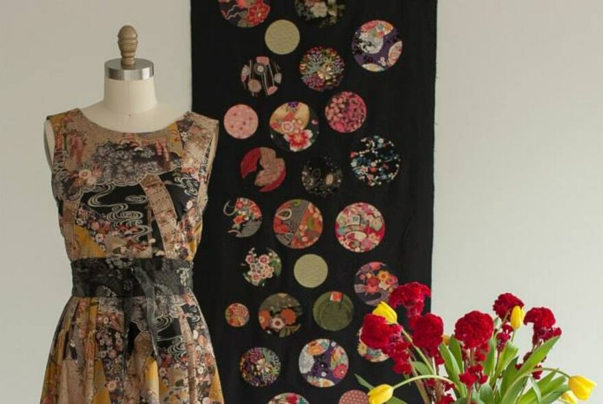 A hand-made dress on a dressmakers' model, with a glass vase of red and yellow tulips beside and a black patchwork hanging behind.