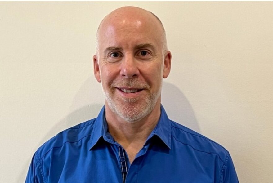 Journalist Michael Bachelard, wearing a blue shirt, smiling slightly and standing in front of a white wall.