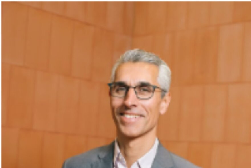 Portrait of a man in a grey suit, wearing glasses, in front of an orange background.