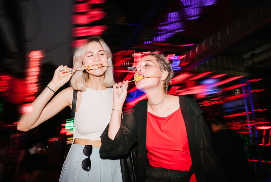 Two young women eating meat off skewers in a neon-lit night scene.