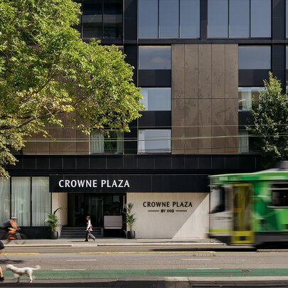 Exterior of Crowne Plaza Melbourne Carlton, a modern hotel featuring a distinct brown facade, a large tree on the footpath and a green tram in motion on the road.