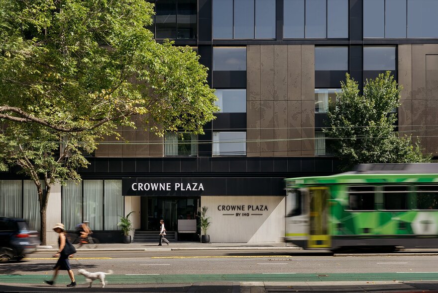 Exterior of Crowne Plaza Melbourne Carlton, a modern hotel featuring a distinct brown facade, a large tree on the footpath and a green tram in motion on the road.