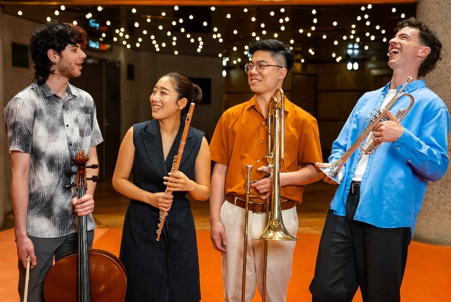 Four musicians holding instruments casually and laughing in a theatre foyer area with fairy lights. 