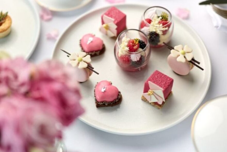 A plate of pink petit-fours arranged neatly and blurred flowers in the foreground. 