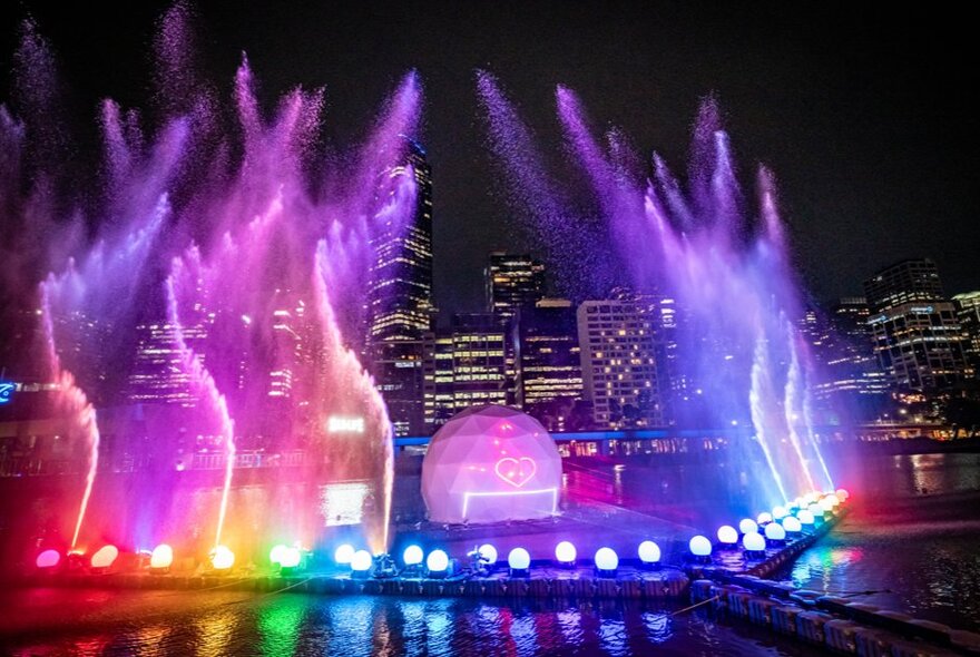 Coloured lights illuminate water sprays high in the air on the Yarra River at night. 