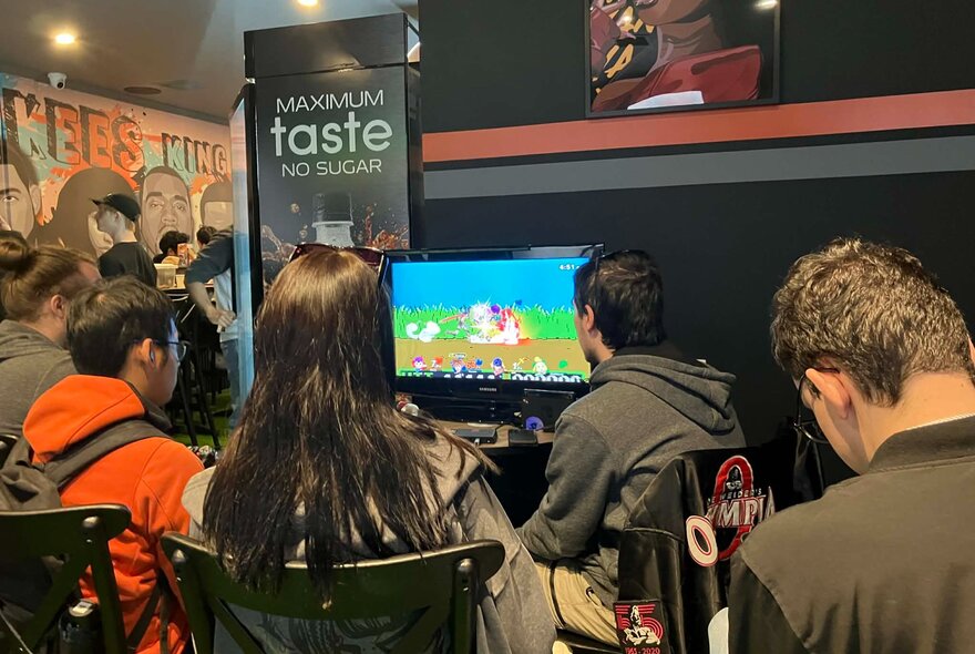 The backs of people's heads seated around gaming consoles inside a cafe or bar.