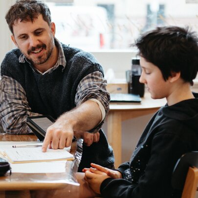 A teacher sits next to a seated student at a table and points at a notepad, during a creative writing workshop in a studio setting.