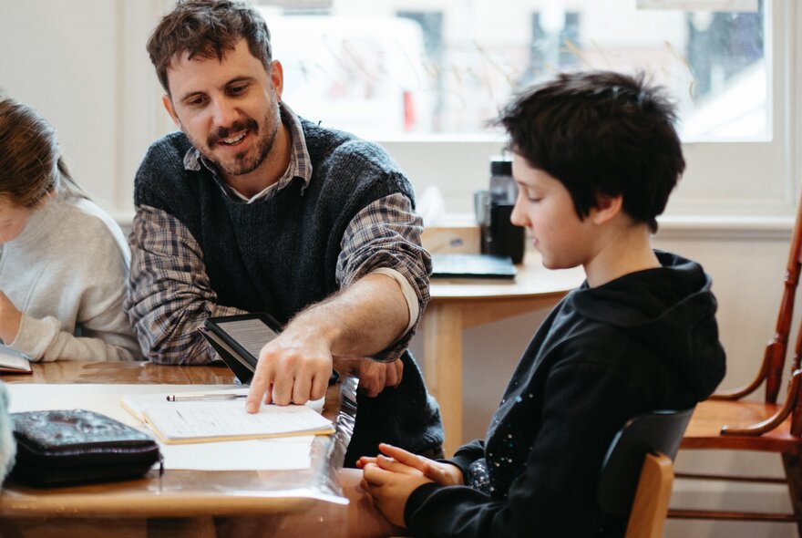 A teacher sits next to a seated student at a table and points at a notepad, during a creative writing workshop in a studio setting.