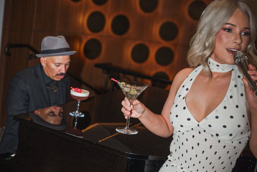 A man seated at a grand piano plays while a woman in a white spotted evening dress sings  while holing a martini.