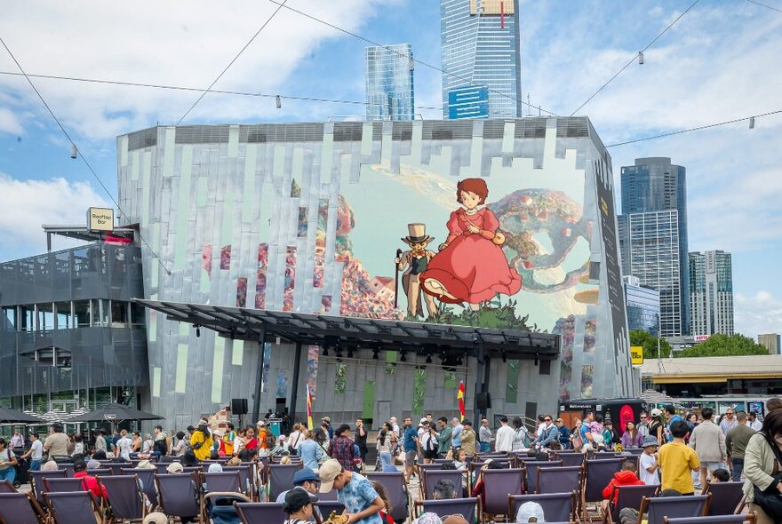 A film showing on the Fed Square big screen, with people watching from deck chairs in the main plaza, with buildings in the background.