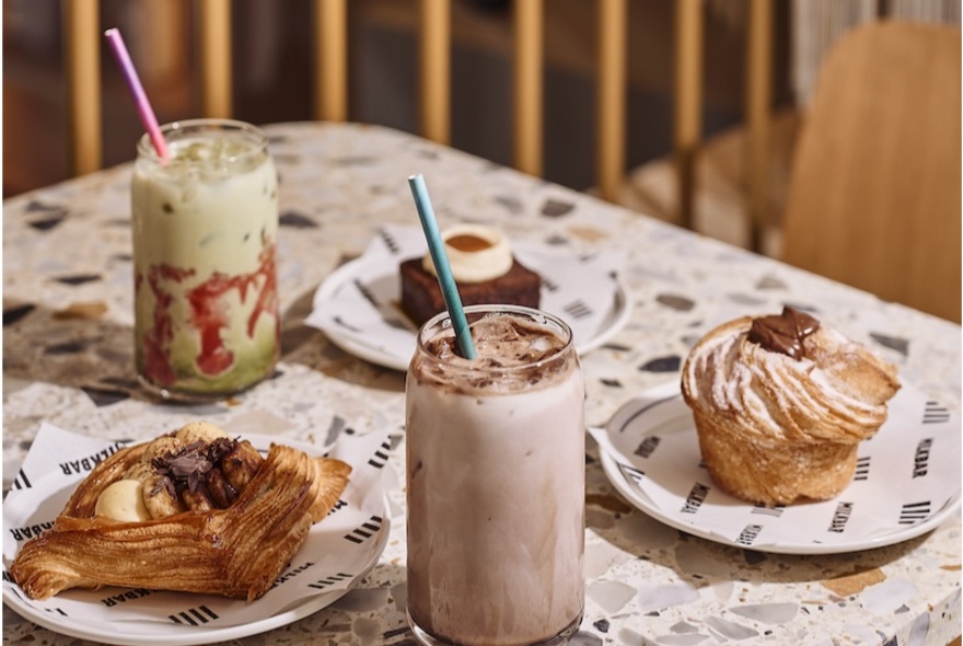 Three pastries on plates, an two iced milk drinks in tall glasses with metal straws, on a terrazzo table top in a cafe.