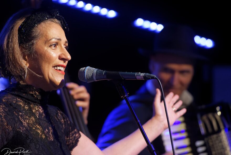 Acclaimed French chanteuse, Noria Letts, singing into a microphone, with a musician playing the accordian behind her.