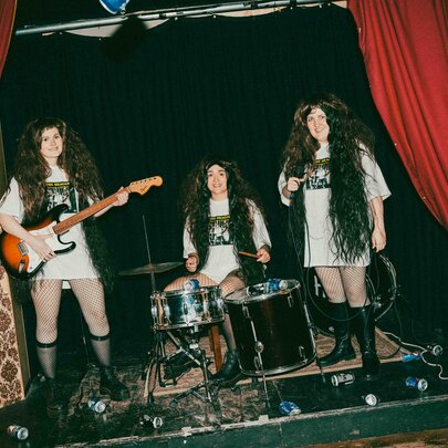 A scene from the theatre show Philosophy of the World with three people on stage, all wearing oversize white t-shirts, and long black hair wigs and playing instruments as if in a band.