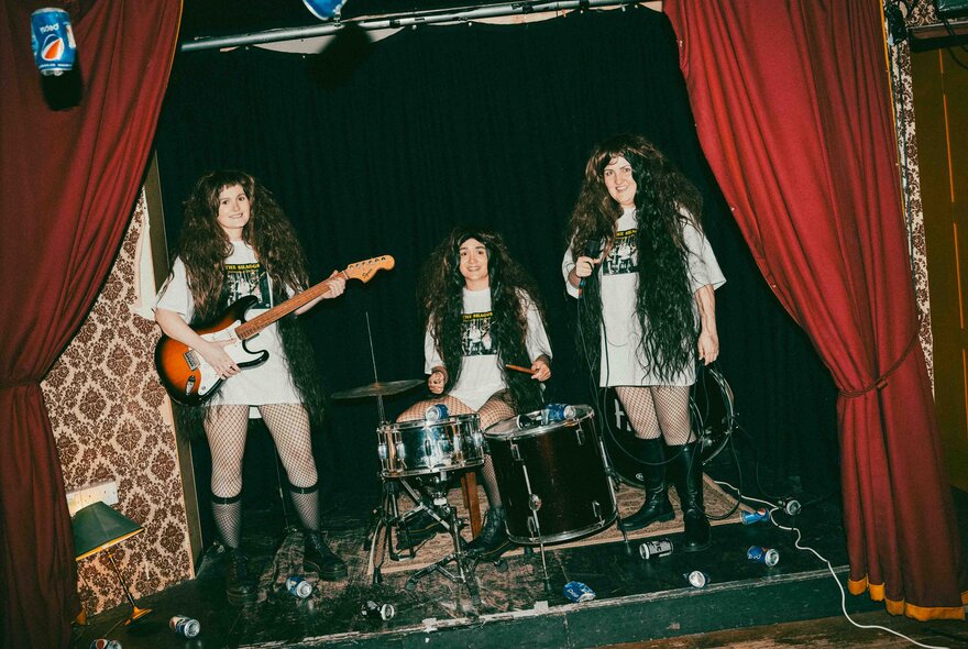 A scene from the theatre show Philosophy of the World with three people on stage, all wearing oversize white t-shirts, and long black hair wigs and playing instruments as if in a band.