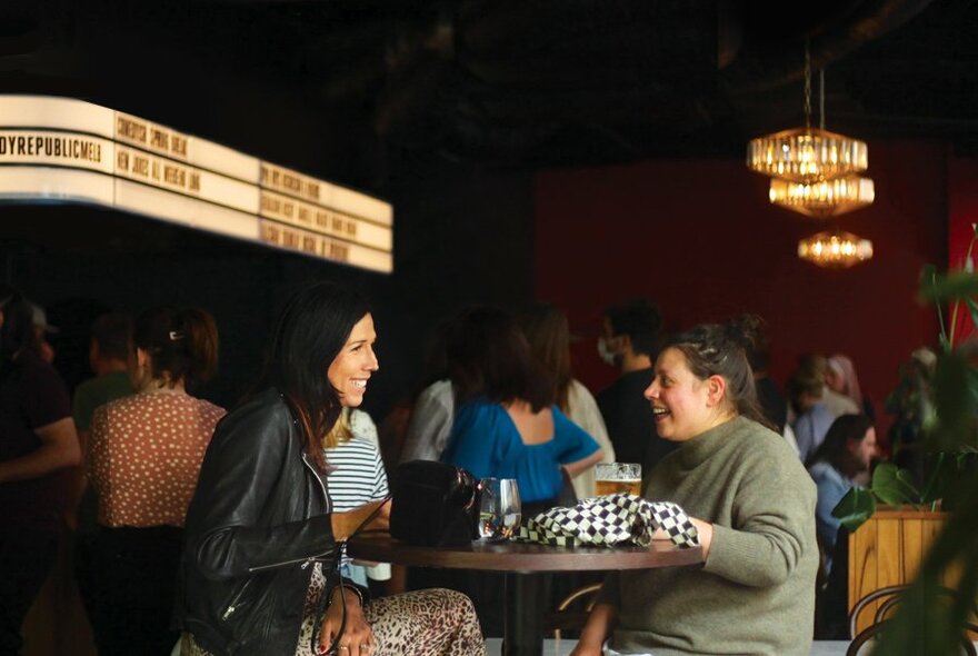 Two women seated in a busy bar, laughing over drinks. 