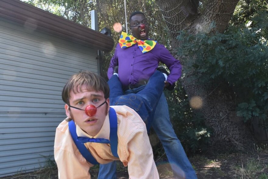 Two performers, one wearing a red clown nose, the other an oversized bright bow tie, holding a wheelbarrow pose outdoors in front of a roller garage door.