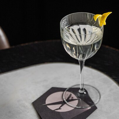 A martini cocktail with a lemon rind garnish resting on the edge of the glass, on a marble-topped table.