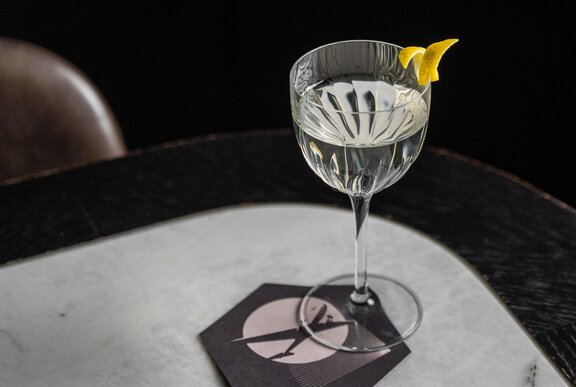 A martini cocktail with a lemon rind garnish resting on the edge of the glass, on a marble-topped table.