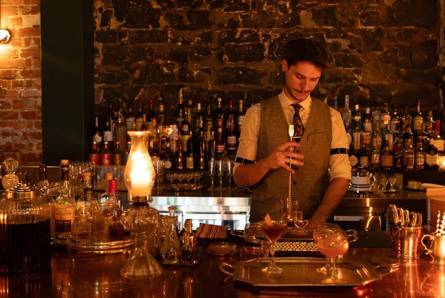 A bartender mixing cocktails at a bar, a wall of bottles and exposed brick behind him.