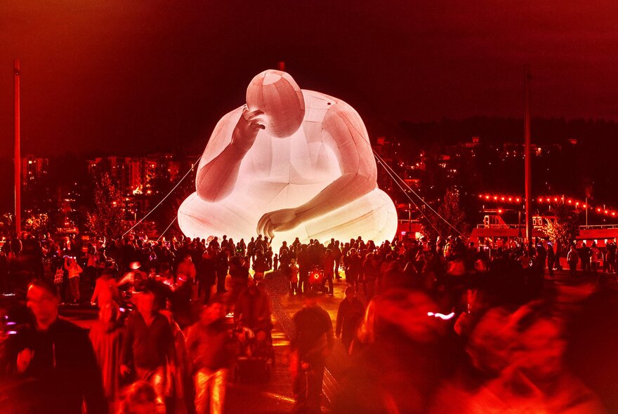 Festival crowds looking at a giant inflatable human sculpture cross legged with its head in its hand.