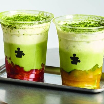 Two flavoured matcha drinks in clear plastic cups on a silver server on a table.
