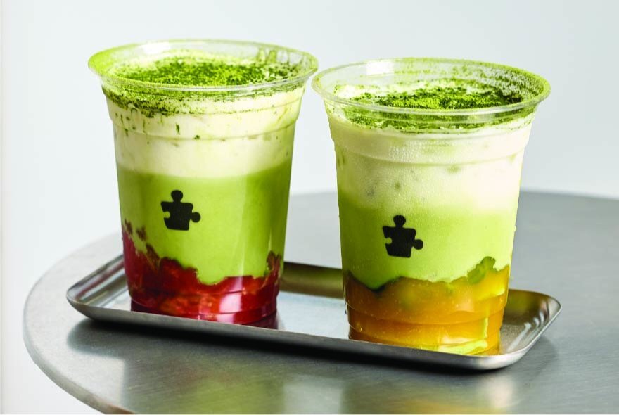 Two flavoured matcha drinks in clear plastic cups on a silver server on a table.