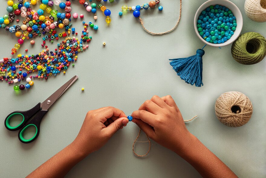Hands threading beads on to a piece of string, with scissors, beads and craft materials.