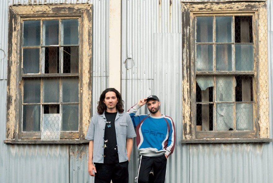 The duo Wax’o Paradiso leaning against the exterior of an old corrugated iron building that has broken windows.