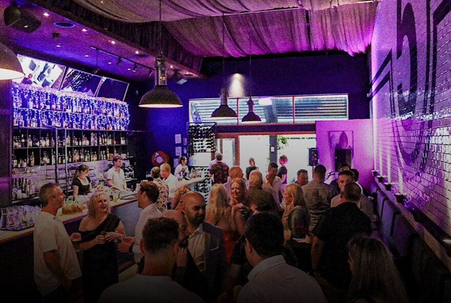 People crowded into a long bar area with purple lights.