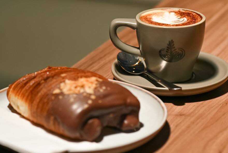 A cup of coffee next to a pastry on a plate on a wooden shelf.