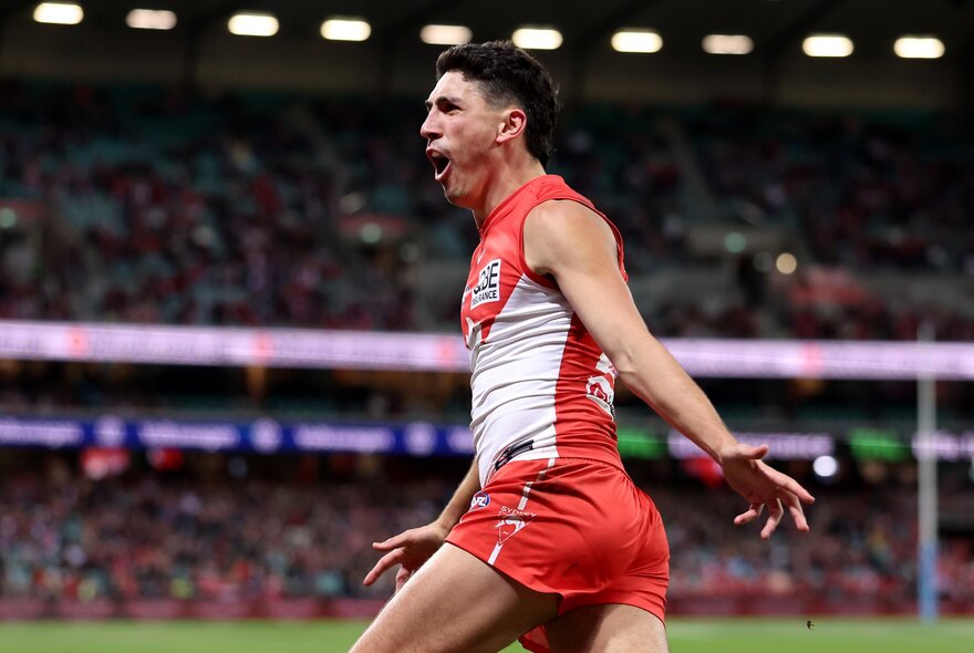 A Sydney Swans AFL player cheering while running on the footy field, blurred fans in the stadium in the background.
