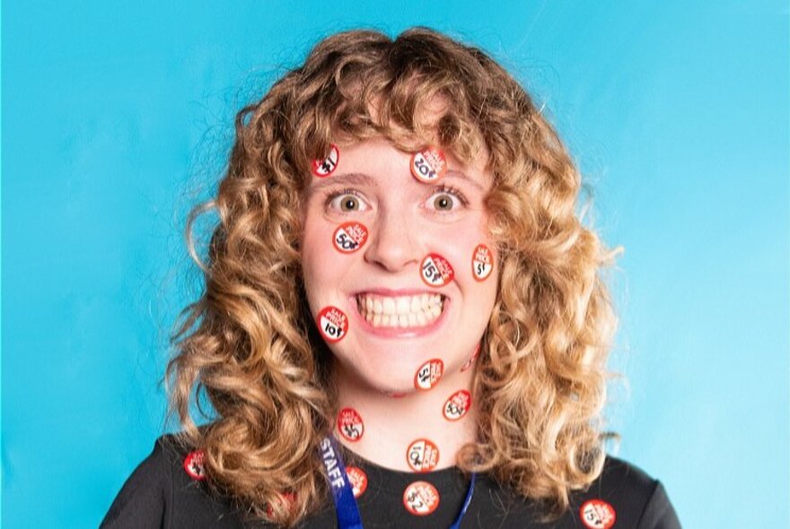 A woman, with curly hair and a forced smile, her face covered in stickers.