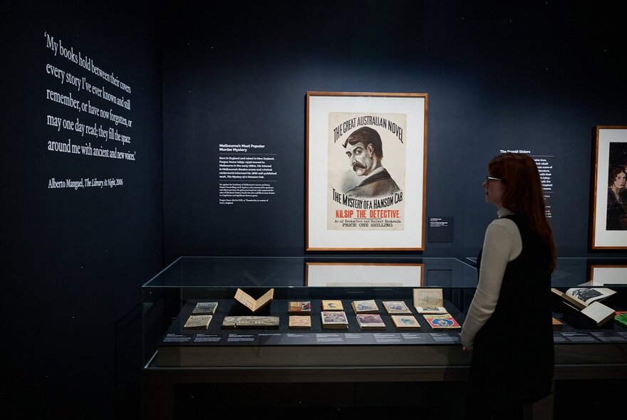 A person at an exhibition in a dark space looking at books in a glass case with a poster behind. 