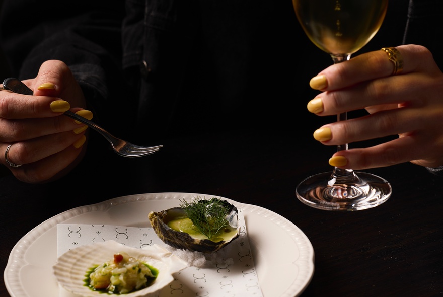 Hands holding a wine glass and fork over a dish of oysters.