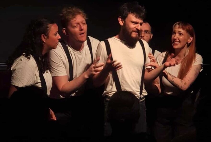 A group of people on stage dressed in white tshirts and braces, gathered around a central man who seems anguished. 