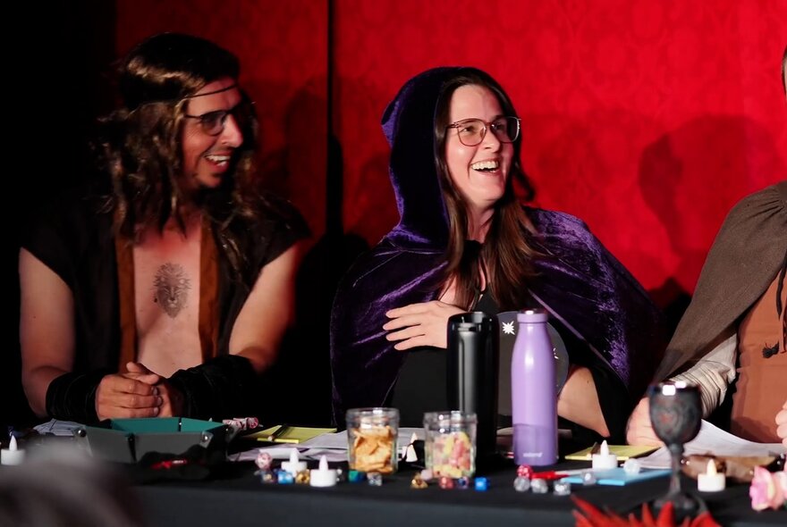 Two performers dressed in character costume, seated at a table on a stage behind a table covered in Dungeons and Dragons props.